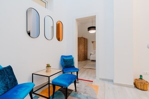 Ein modernes Wohnzimmer mit einer Couch, einem kleinen Tisch und einem blauen Sessel. An der Wand befinden sich drei Spiegel, eine Hängeleuchte und im Hintergrund ein Holzschrank.