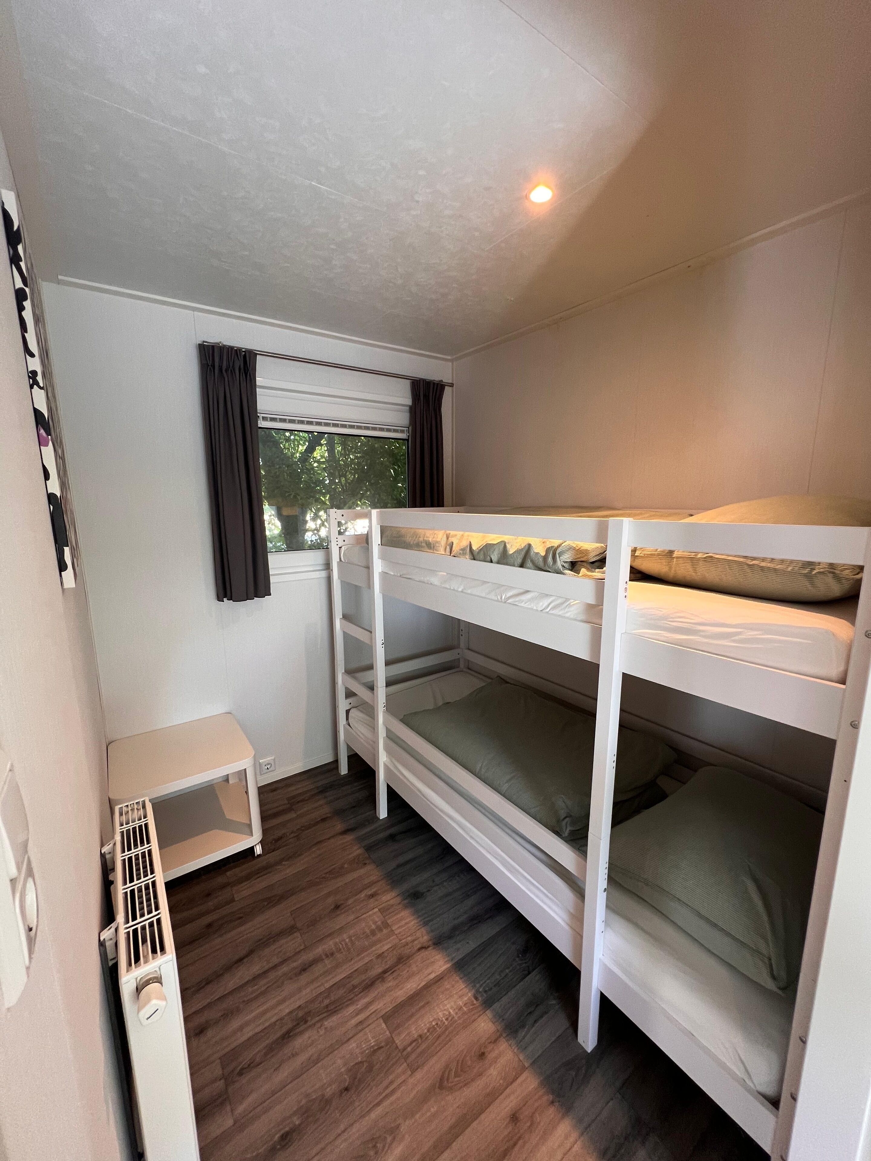 Een kleine, goed verlichte kamer met een stapelbed, een klein tafeltje en een raam met gordijnen.