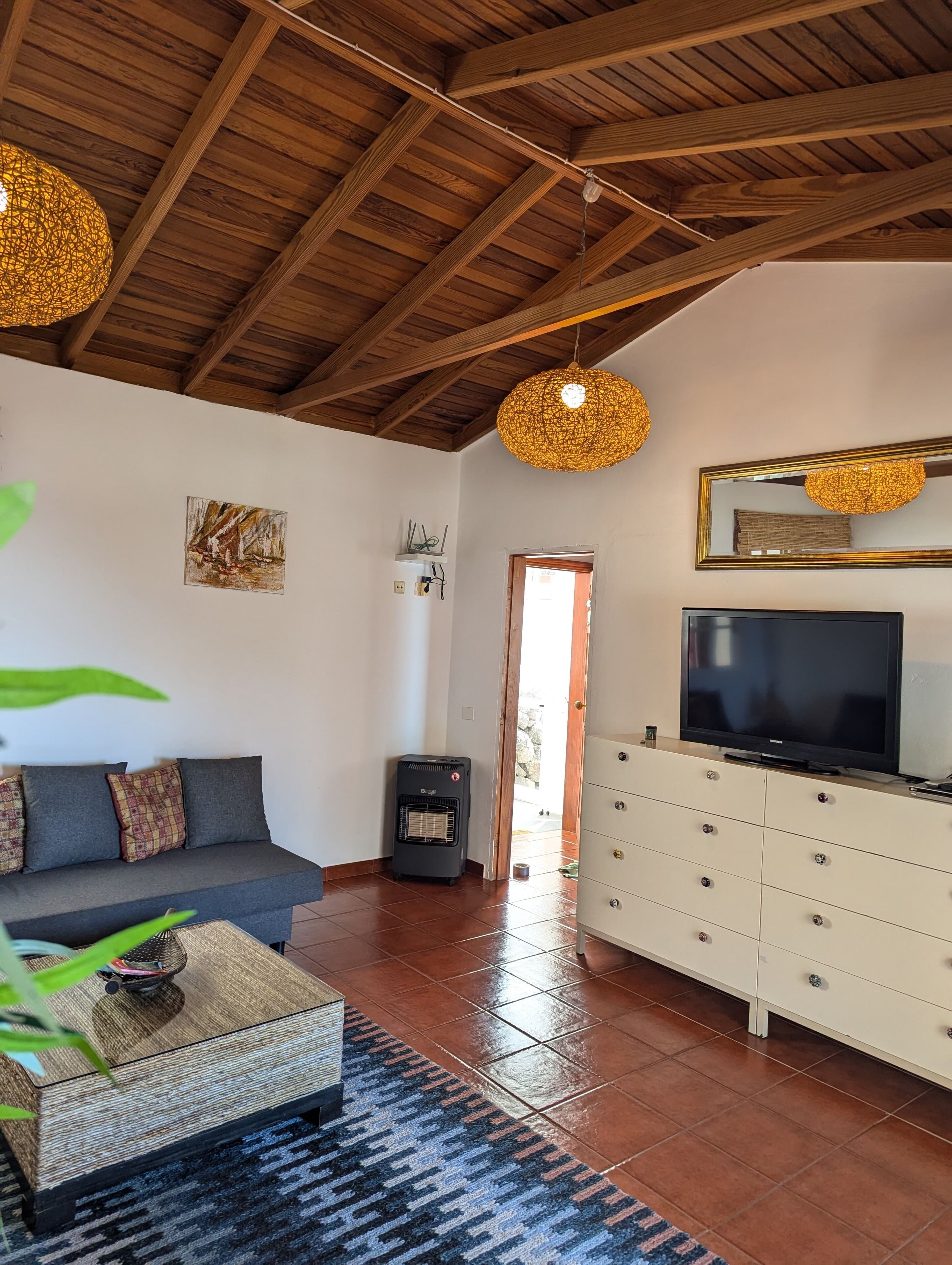 A living room with a wooden ceiling, a sofa, a TV on a cabinet, and a door leading outside.
