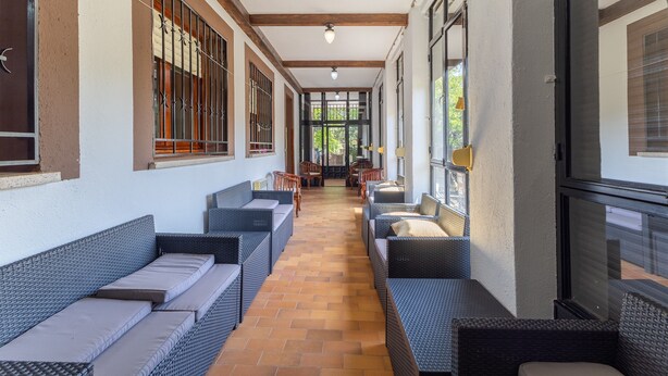 Un corredor techado con muebles de mimbre, ventanas amplias y suelo de baldosas.