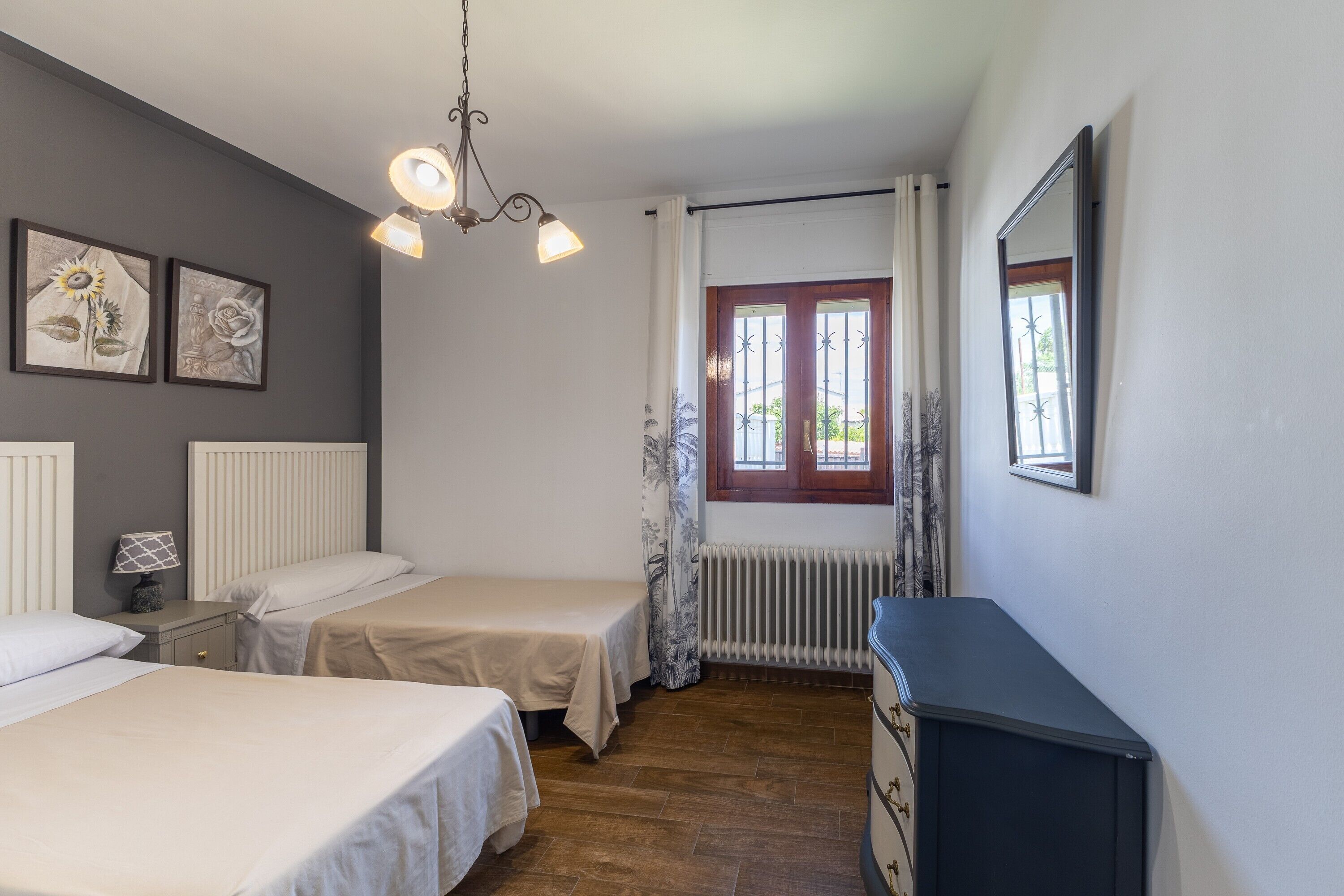 Un dormitorio con dos camas, un aparador, un radiador, una ventana con cortinas y obras de arte colgadas en la pared.