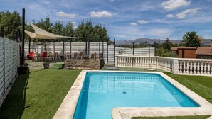 Piscina rectangular con un deck blanco, rodeada por una valla blanca y una zona de sombra con sillas.