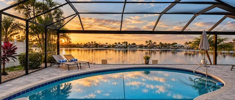A swimming pool with a view of a sunset over water, surrounded by lounge chairs and palm trees.