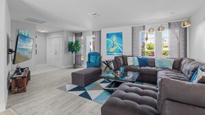 A modern living room with a sectional sofa, a coffee table, and a TV mounted on the wall. There are two chairs, a potted plant, and artwork on the walls. - Intervillas Florida - Villa Bluesky at the Lake (Cape Coral)