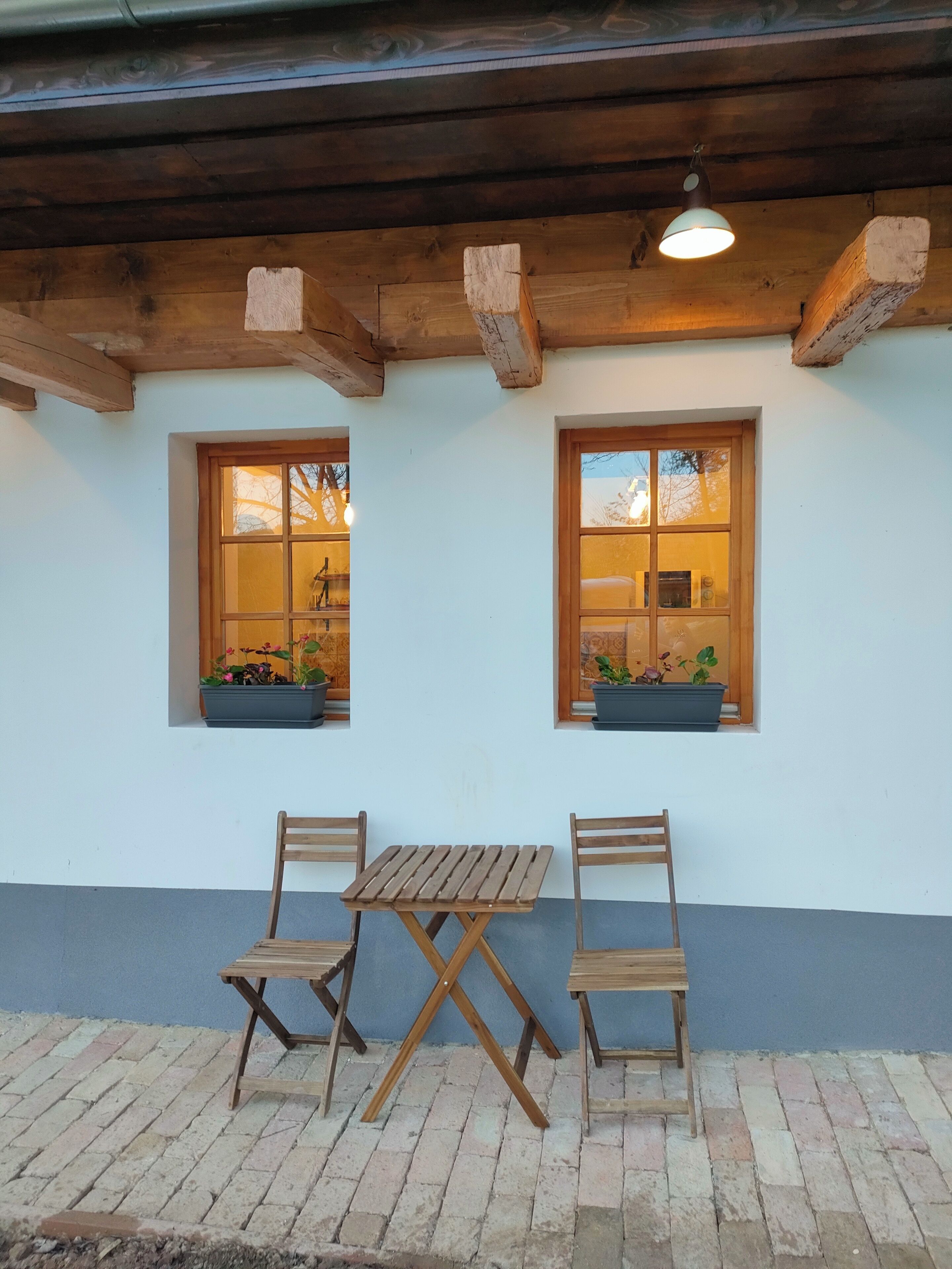 A wooden table and two chairs under a wooden beam ceiling with two windows, each with a plant.