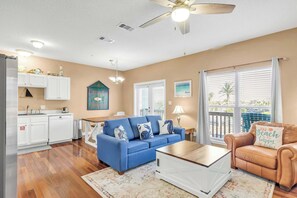 A living room with a blue sofa, a brown leather chair, and a wooden coffee table. There is a kitchen area with white cabinets and a refrigerator. A ceiling fan is visible, and the room has large windows with blinds. - Barkentine: Pool & White Sands Steps Away (Fort Walton Beach)