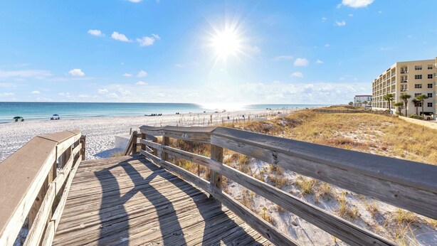 Une promenade en bois qui mène à une plage, avec un ciel dégagé et un soleil éclatant.