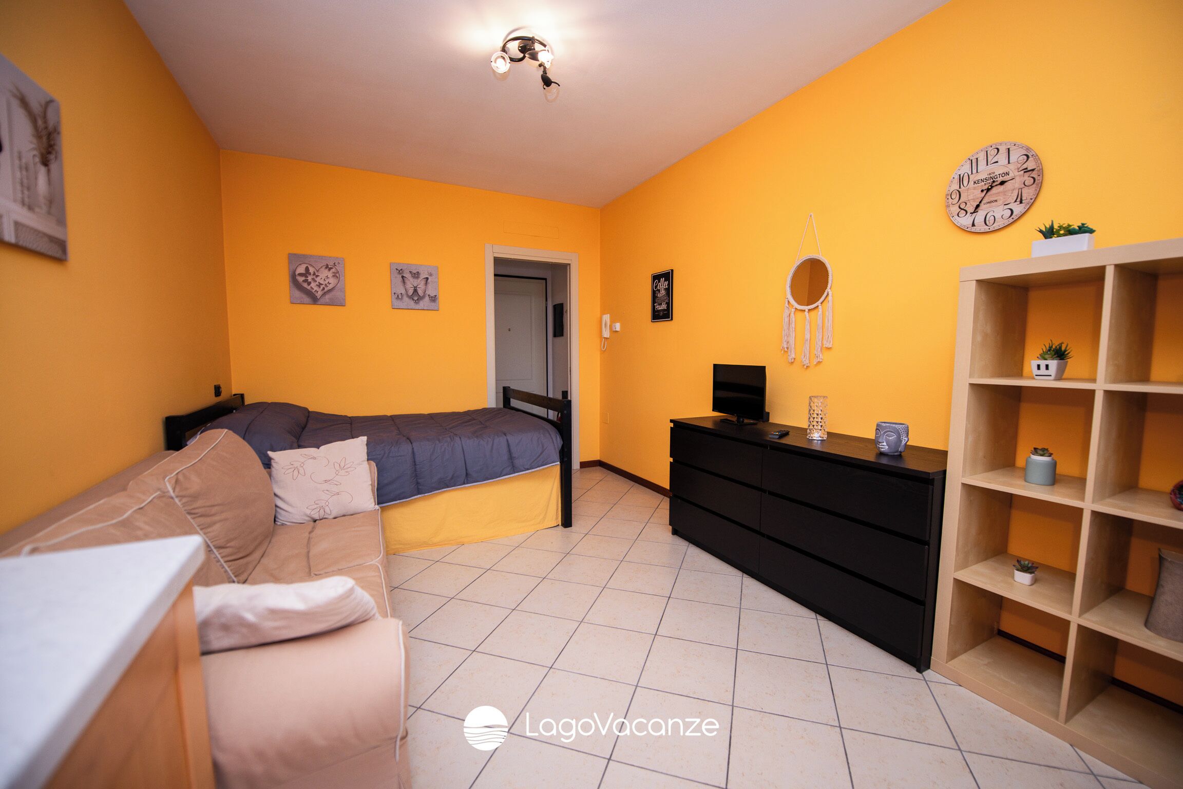 Een slaapkamer met een bed, een dressoir, een plank met decoratieve objecten en een wandklok.