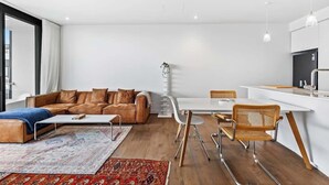 Een moderne woonruimte met een bruine leren bank, een witte eettafel met stoelen, en een keuken met witte kasten en een zwart aanrecht.