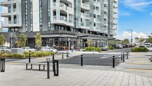 Een modern gebouw met een glazen gevel, een overdekte ingang en een parkeerplaats met auto's en bankjes.