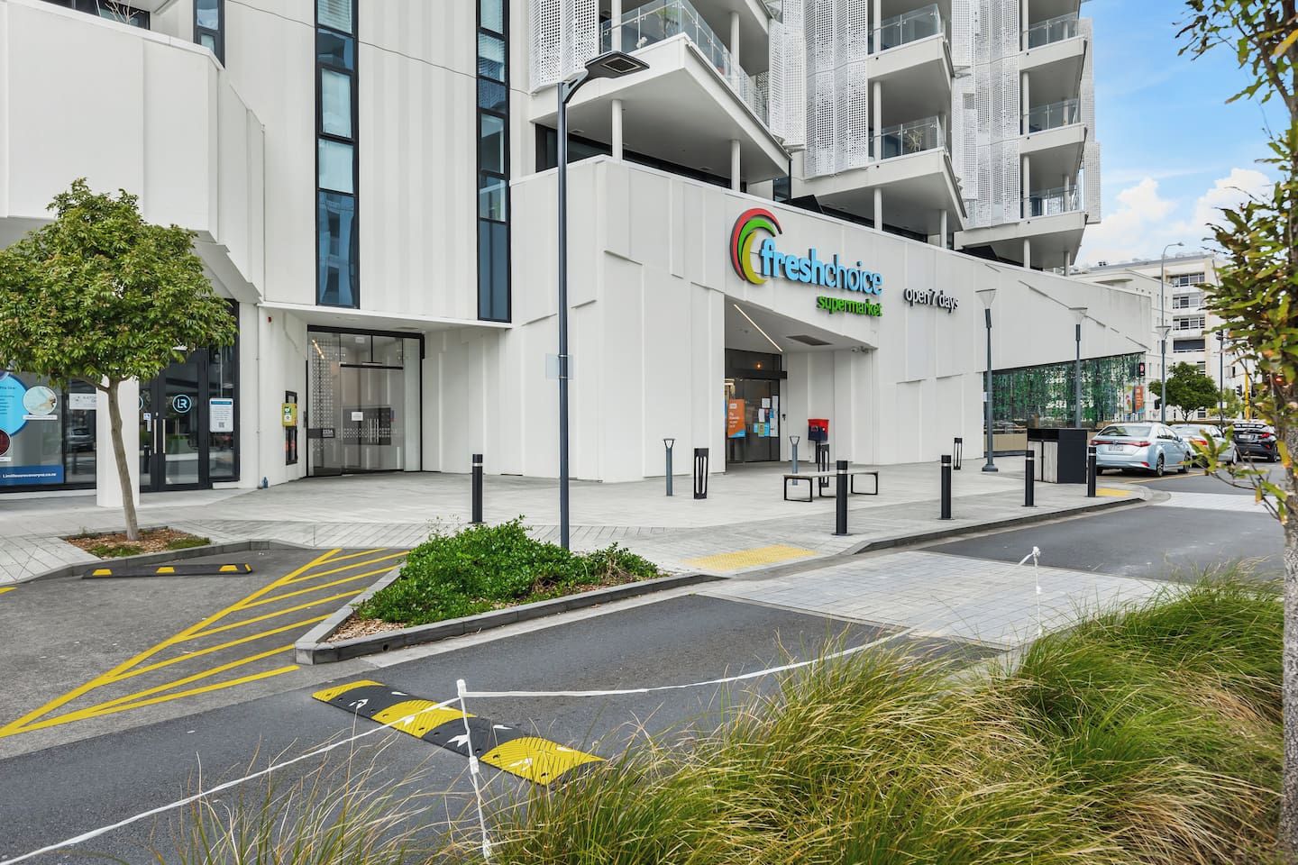 A modern building with a FreshChoice supermarket entrance, a clear pedestrian pathway, and a parking area with marked spaces.