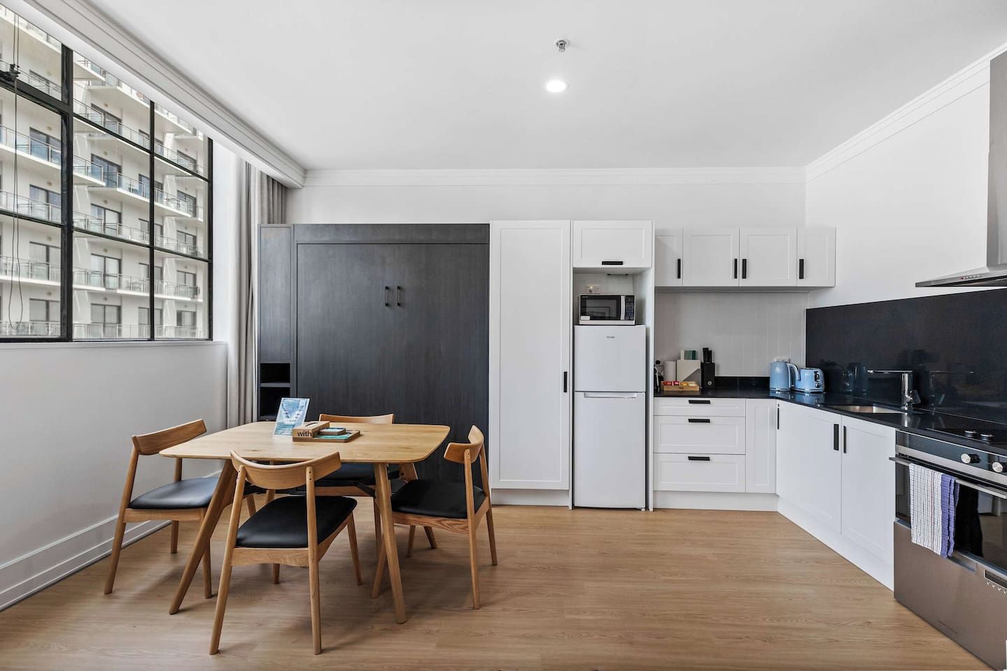 A modern kitchen with a dining area, white cabinetry, a black countertop, and a view of a building through large windows.