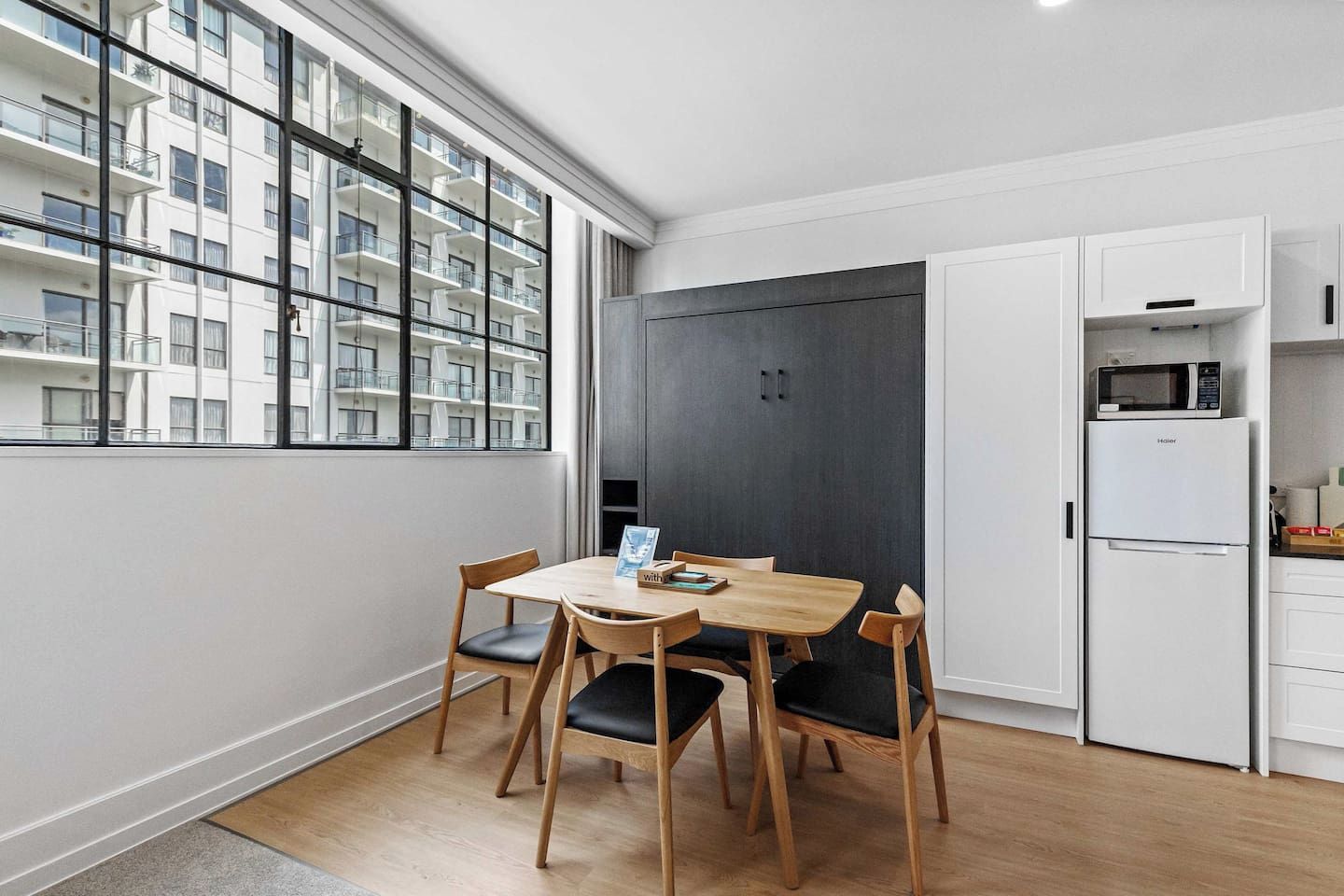 A modern kitchen with a dining table, chairs, a refrigerator, and a view of a building through large windows.