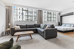 Ein modernes Wohnzimmer mit einem Ecksofa, einem Bett, einem kleinen Tisch und großen Fenstern mit Blick auf Gebäude.