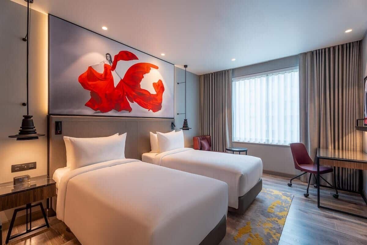 A hotel room with two beds, a desk, a chair, and a large abstract painting on the wall.