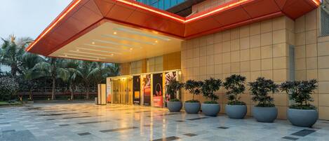 Una entrada de hotel moderna con fachada de vidrio geométrico, plantas en macetas grandes y una plaza adoquinada.