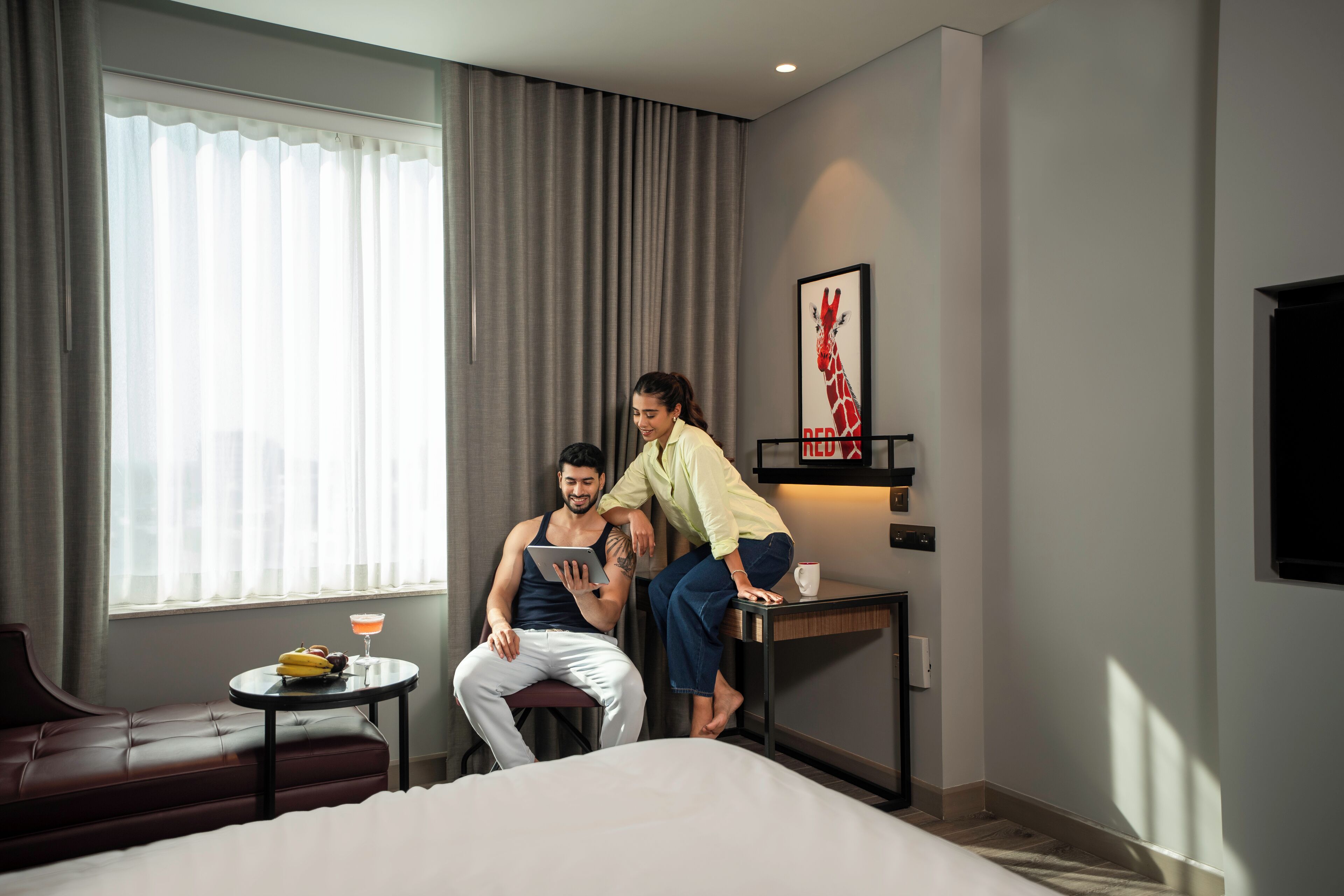 A hotel room with a bed, a small table with a drink, a sofa, and a large window with curtains.