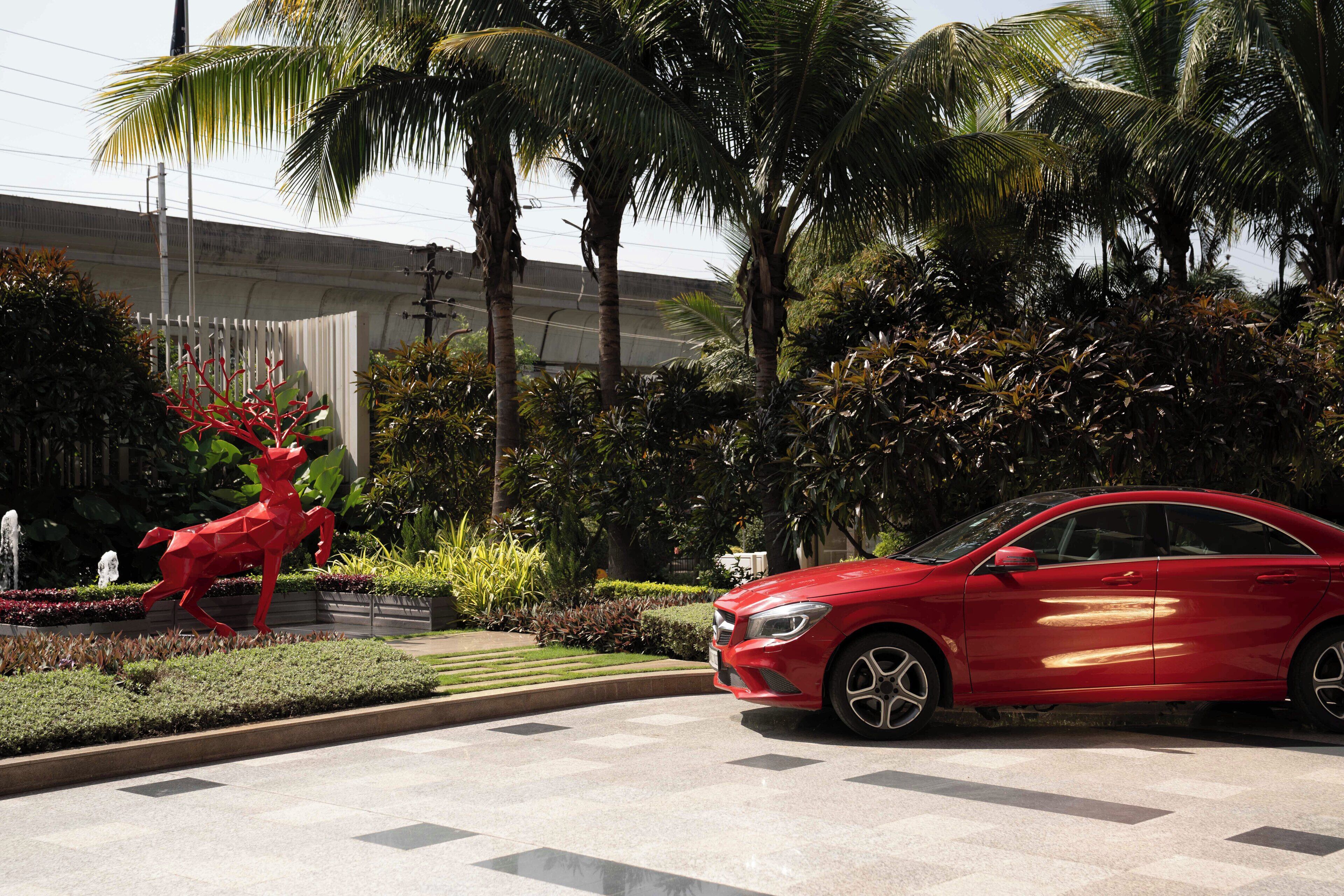 A red car parked near a sculpture of a deer in a landscaped area with palm trees and a building in the background.