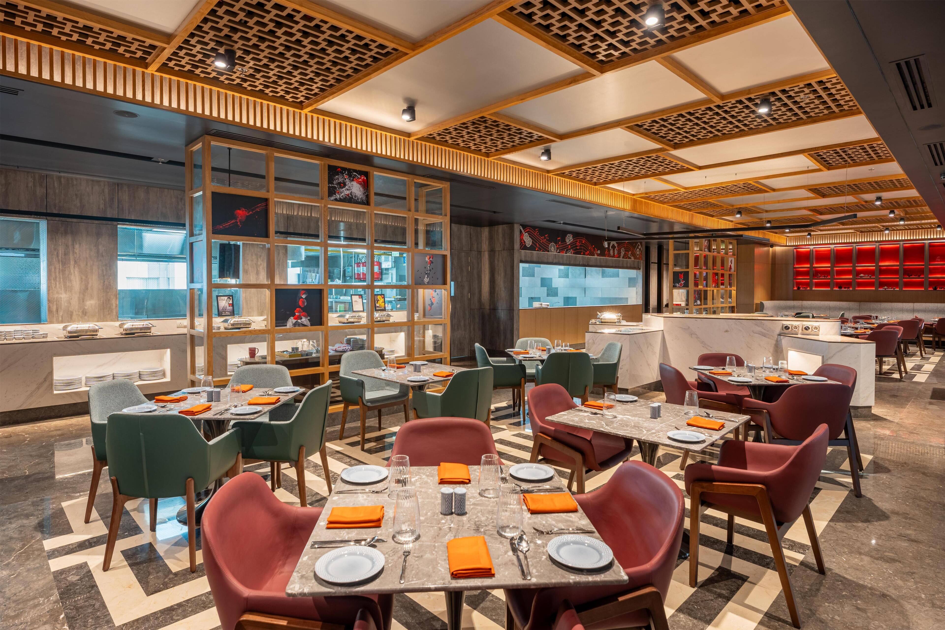 A modern restaurant with a central bar area, multiple dining tables, and a mix of green and red chairs.