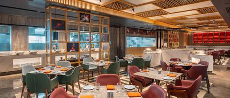 A modern restaurant with a central bar area, multiple dining tables, and a mix of green and red chairs.