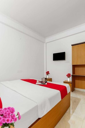 A hotel room with a bed, a TV mounted on the wall, a wooden wardrobe, and a small table with a vase of flowers. - Hotel Awadh Pearl (Lucknow)