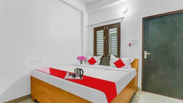 A neatly made bed with a red and white bedspread, a wooden headboard, and a small table with a kettle and a vase of flowers. - Hotel Awadh Pearl (Lucknow)