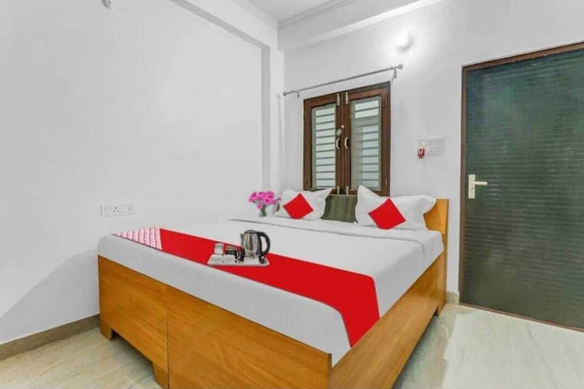 A neatly made bed with a red and white bedspread, a wooden headboard, and a small table with a kettle and a vase of flowers.