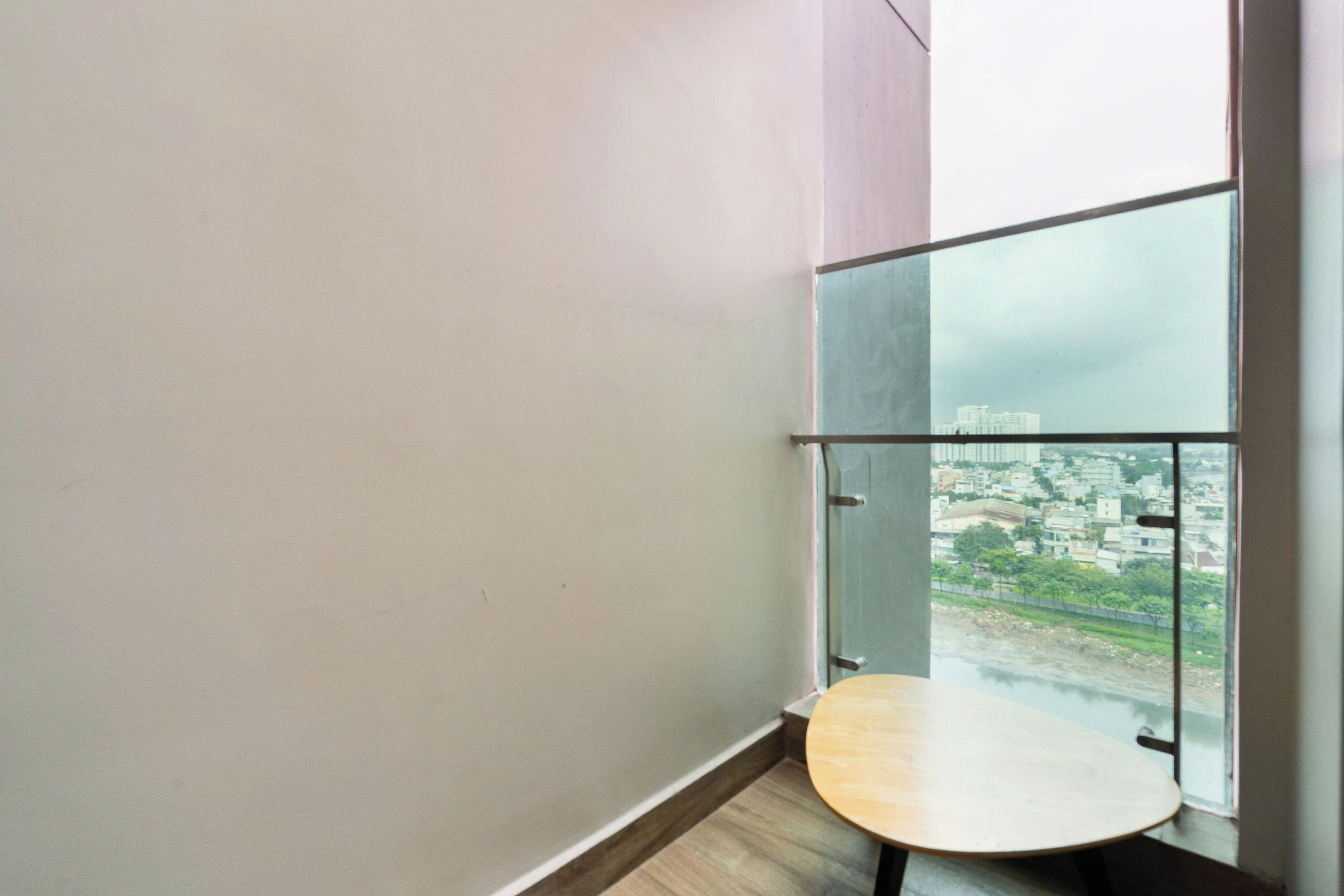 A small balcony with a glass railing, a wooden table, and a view of a cityscape through the window.