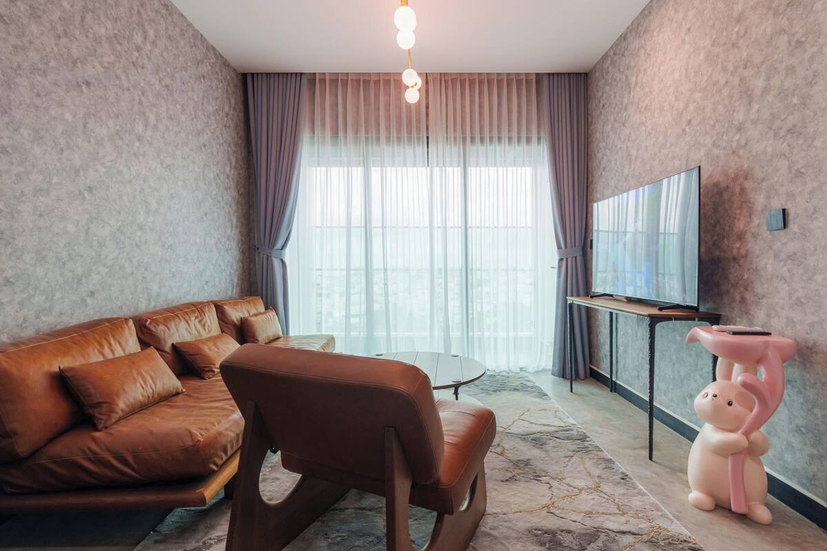 Ein modernes Wohnzimmer mit einem braunen Ledersofa, einem kleinen runden Tisch, einem pinkfarbenen Schwein-Sitzhocker, einem Fernseher auf einem Schrank und einem großen Fenster mit Vorhängen.