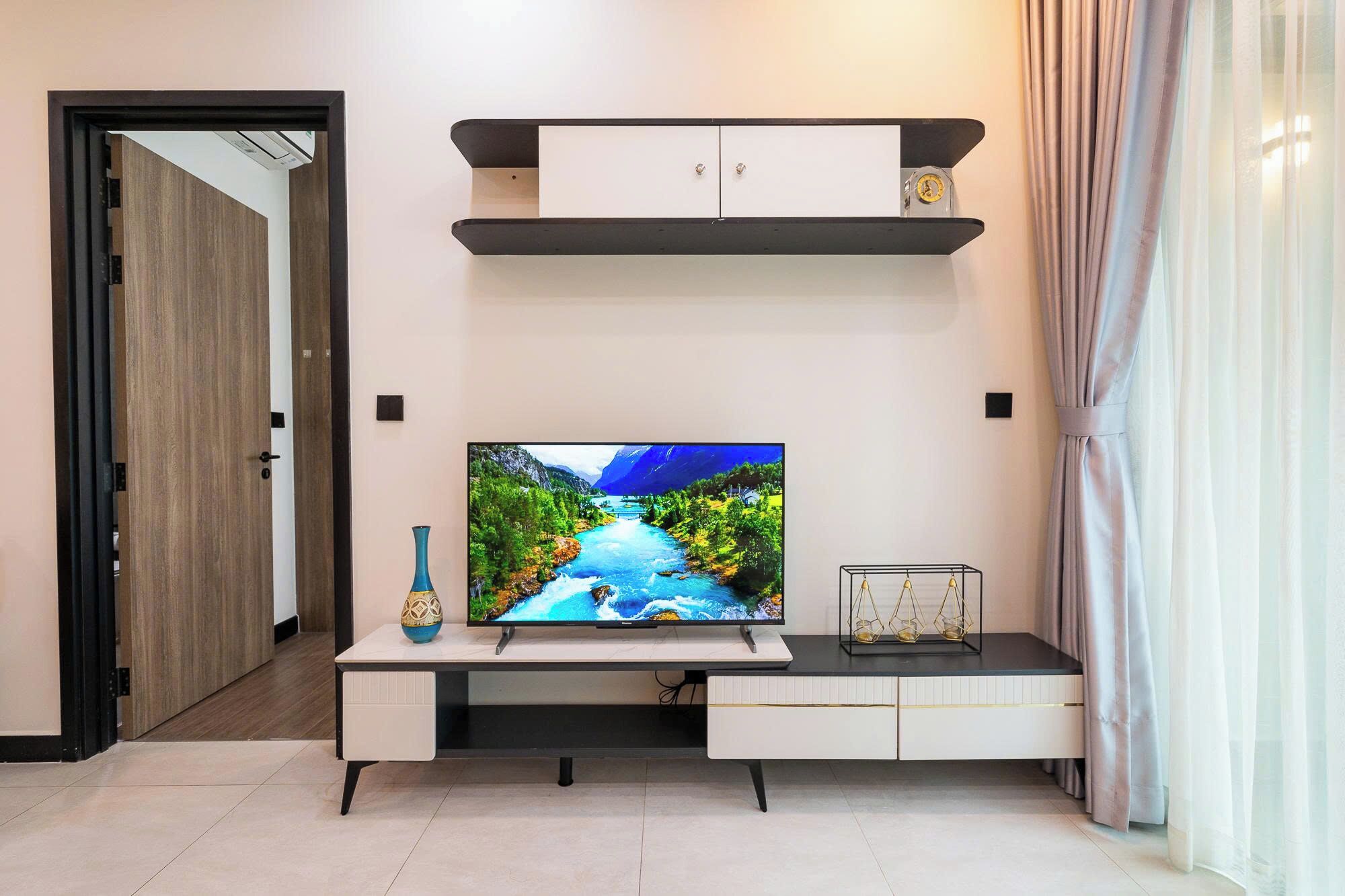 Ein modernes Wohnzimmer mit einem Flachbildfernseher auf einem weißen TV-Schrank, einer dekorativen Vase und einem schlichten Regal darüber.