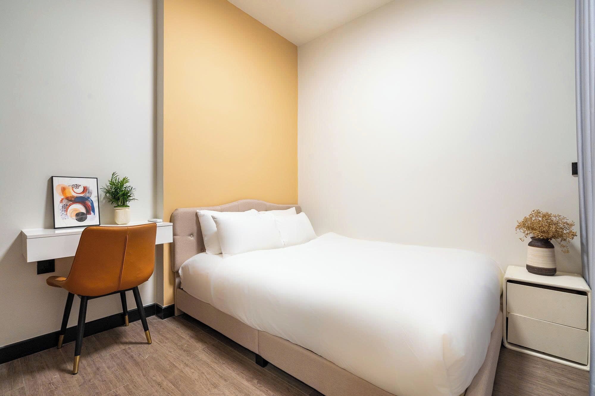 A hotel room with a bed, a desk, a chair, a small table with a vase, and a plant.