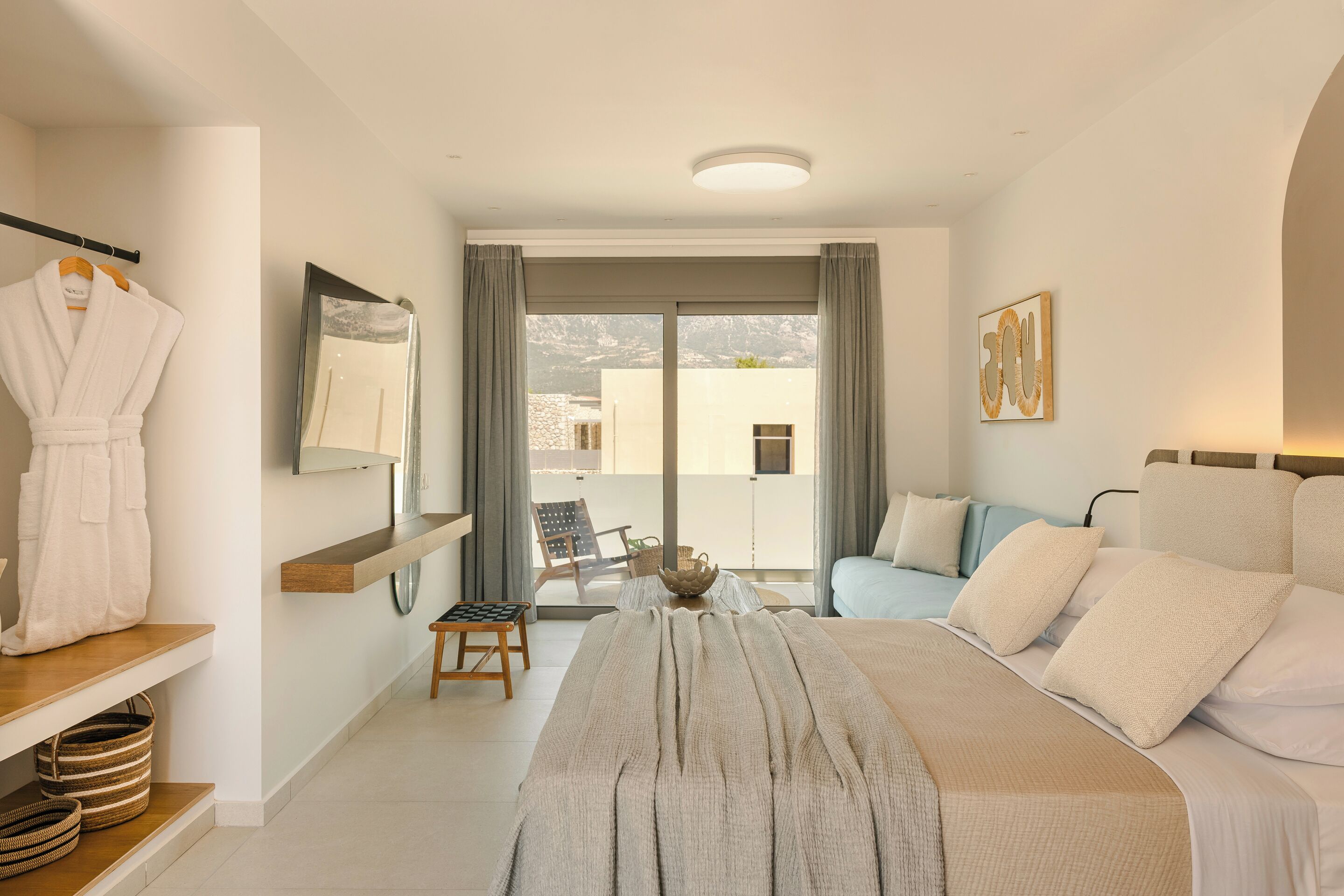A modern hotel room with a bed, a seating area with a table, a robe hanging, and a view of the outside.