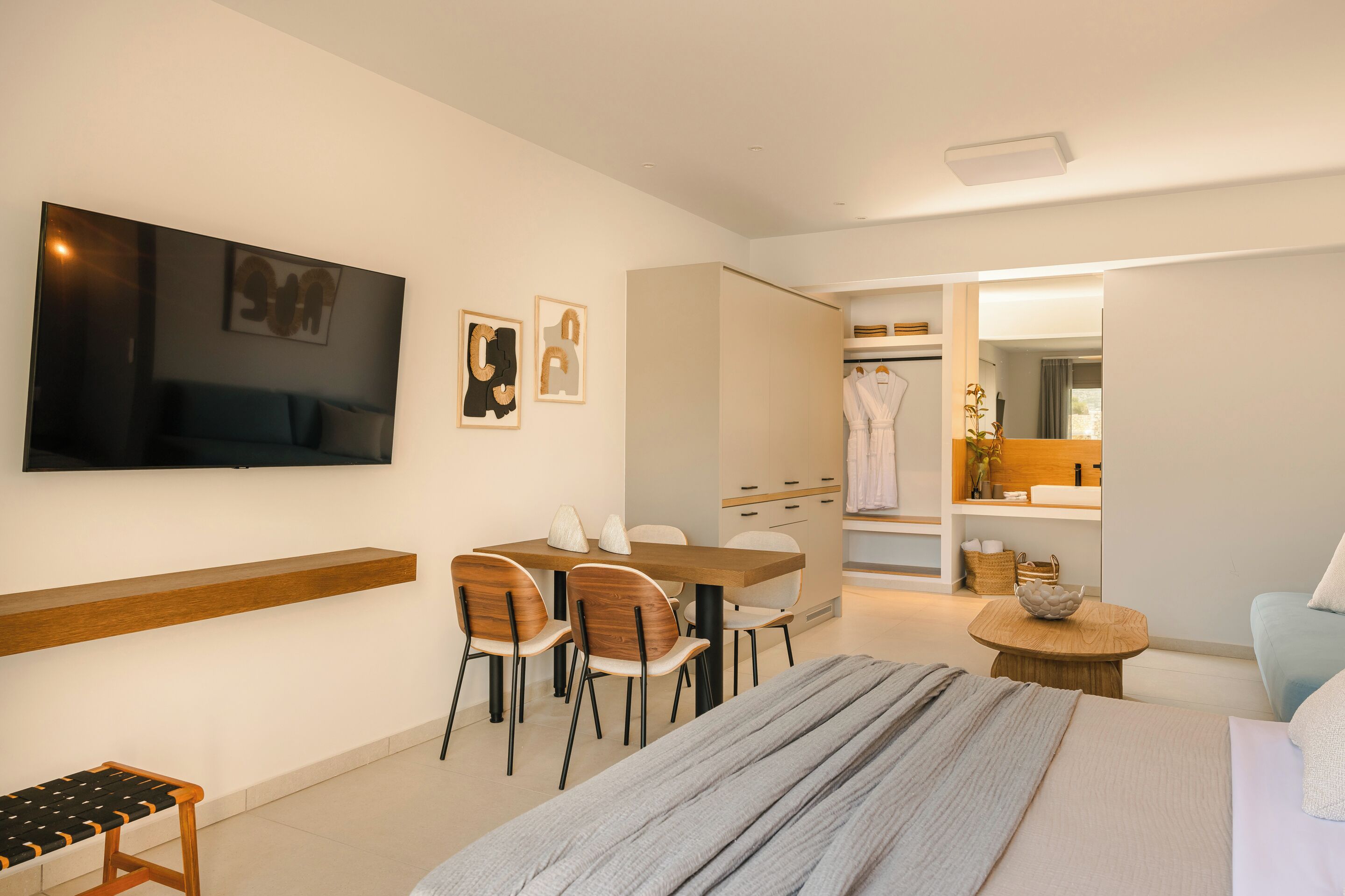 Una habitación de hotel moderna con una cama, un televisor montado en la pared, una zona de comedor con sillas y una mesa, y un baño visible a través de una puerta abierta.
