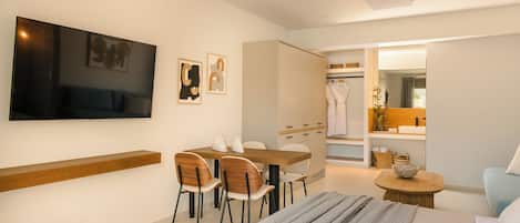 Una habitación de hotel moderna con una cama, un televisor montado en la pared, una zona de comedor con sillas y una mesa, y un baño visible a través de una puerta abierta.