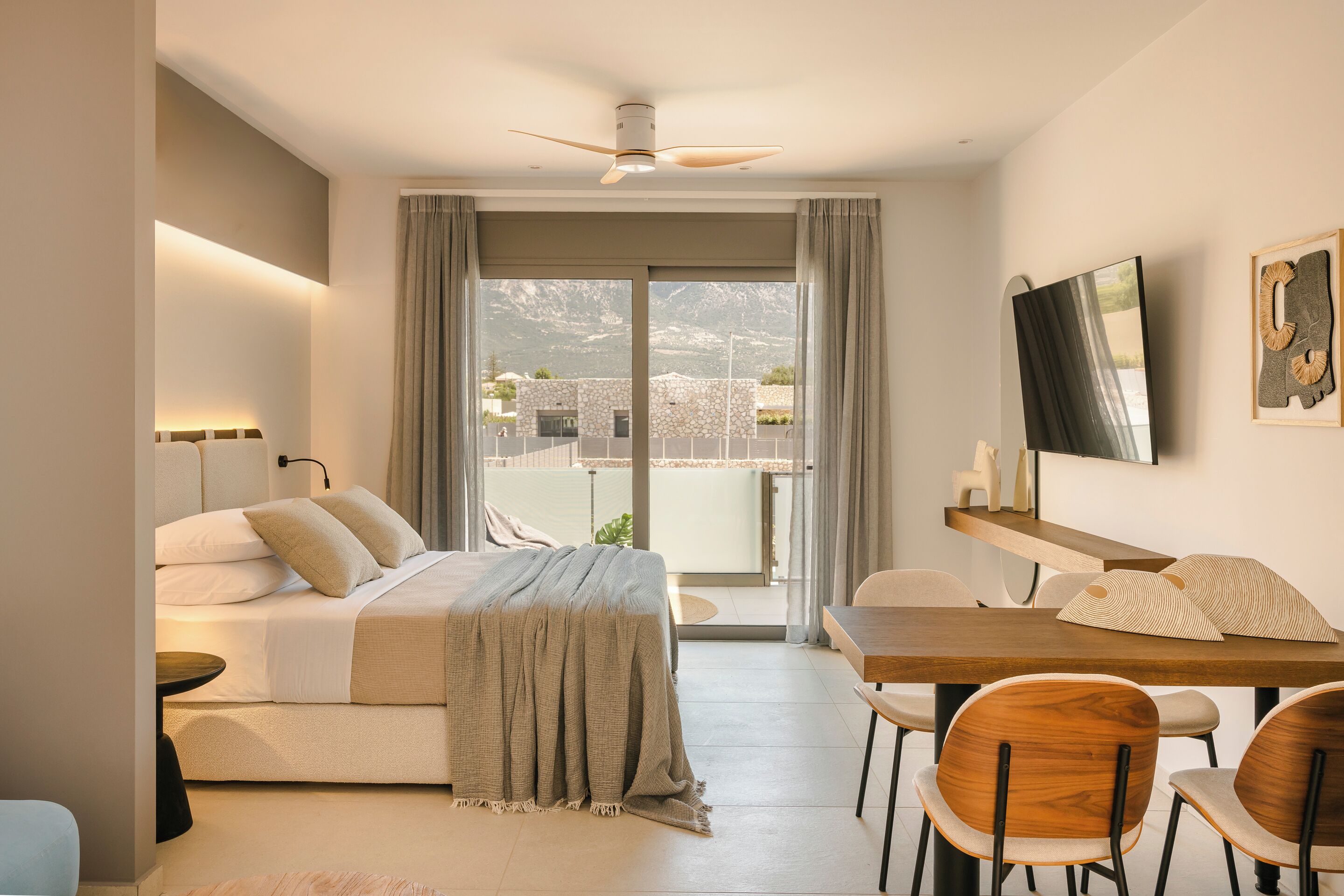 A modern hotel room with a bed, a dining table, and chairs. There is a balcony with a view of mountains and a building.