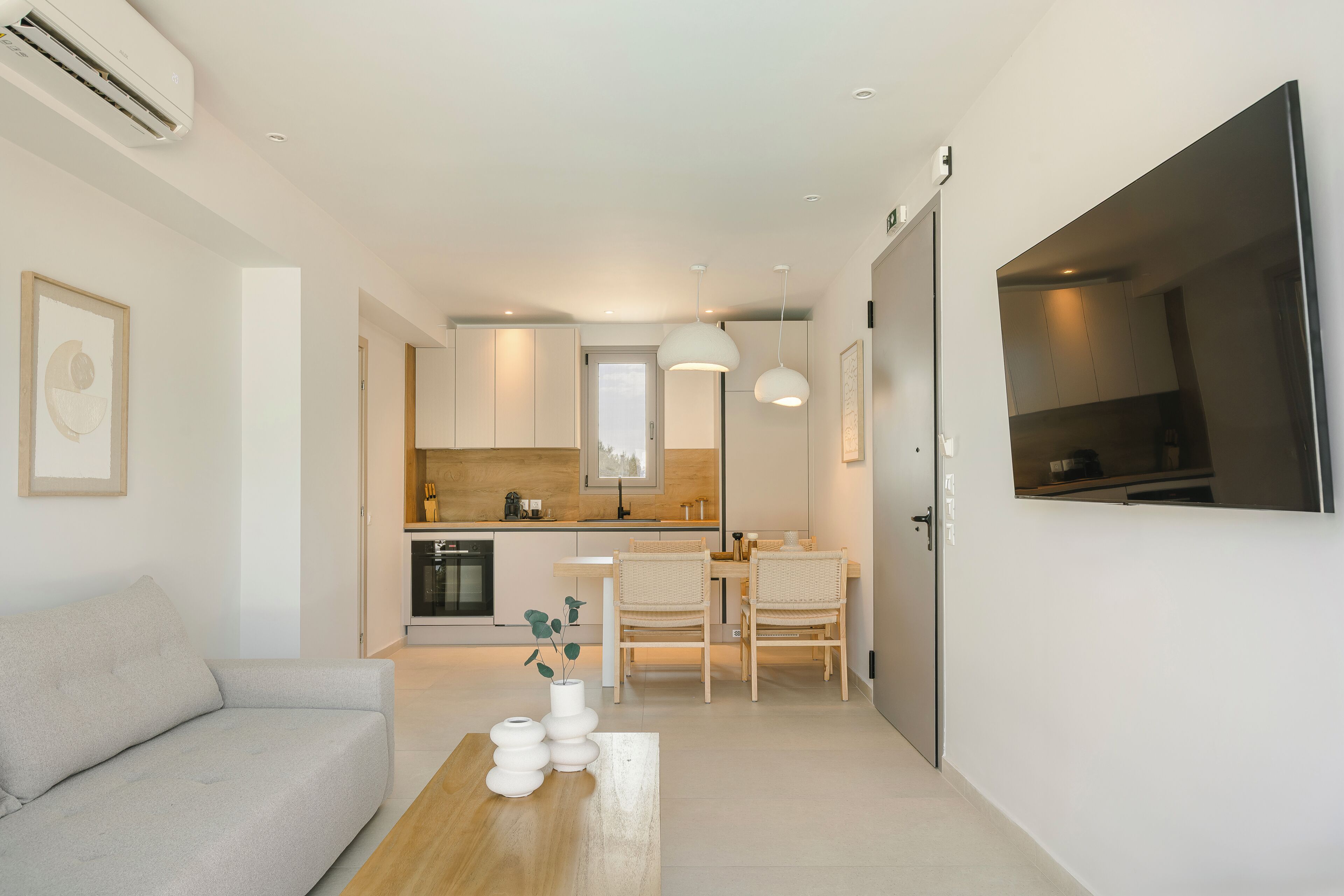 A modern living space with a sofa, dining table, and chairs. A kitchen is visible in the background with a large window.