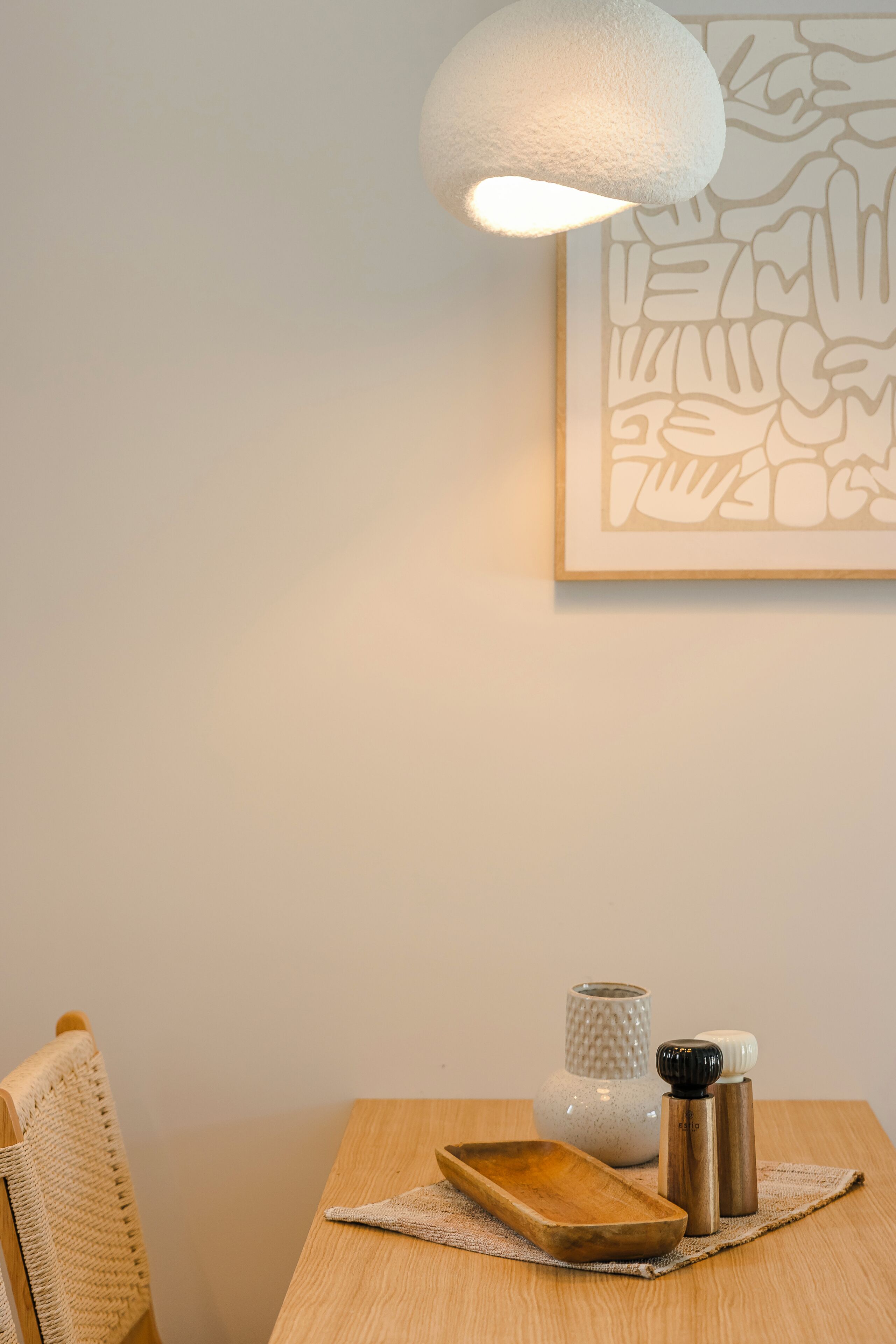 A wooden table with a white textured lamp, a framed abstract art piece, a white vase, and two metallic salt and pepper shakers.