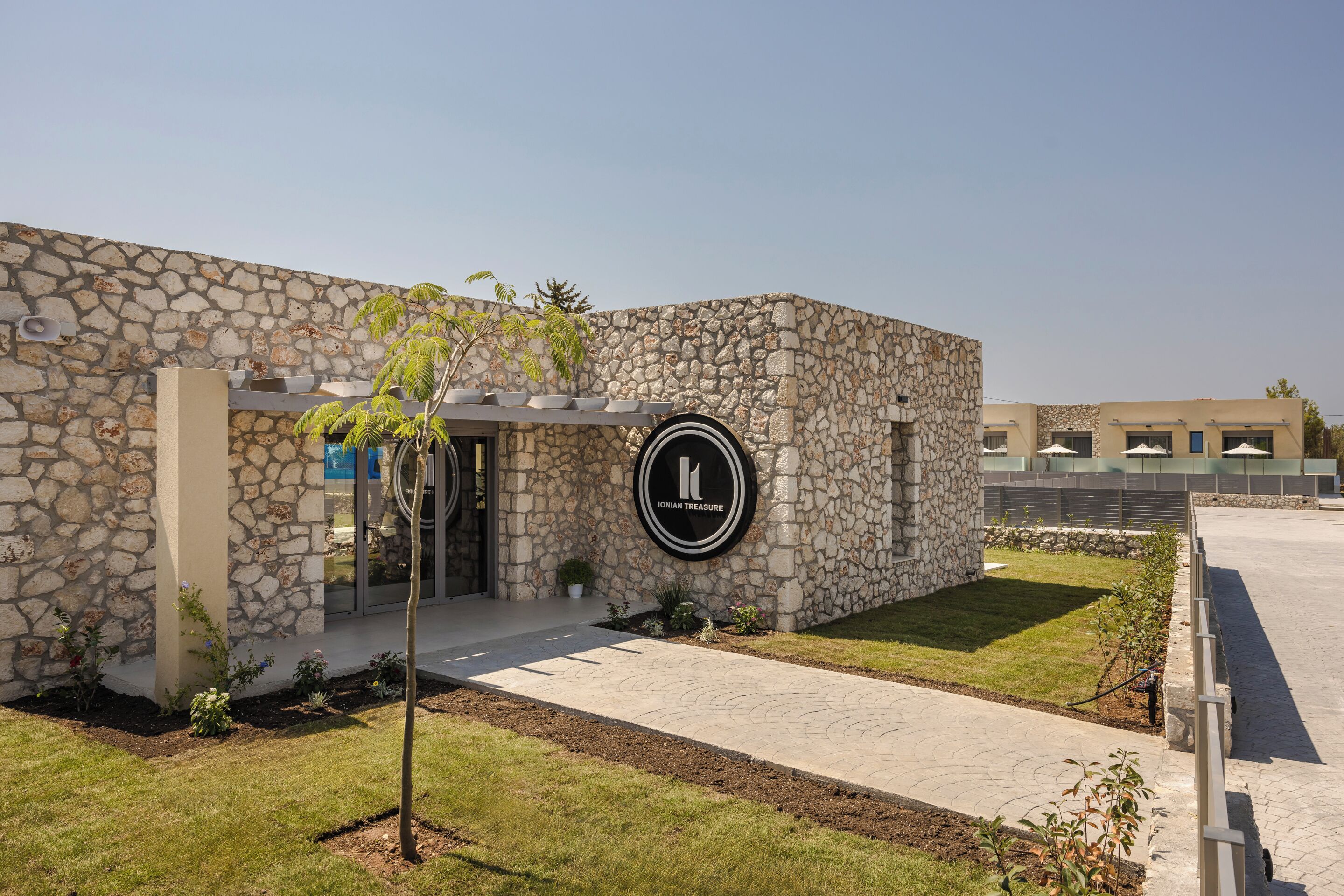 A stone building with a circular logo, a paved entrance, and a small garden area.
