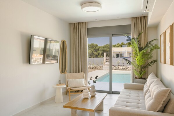 A modern living room with a sofa, a coffee table, a chair, a TV, and a view of a swimming pool. - Ionian Treasure Suites (Kefalonia)