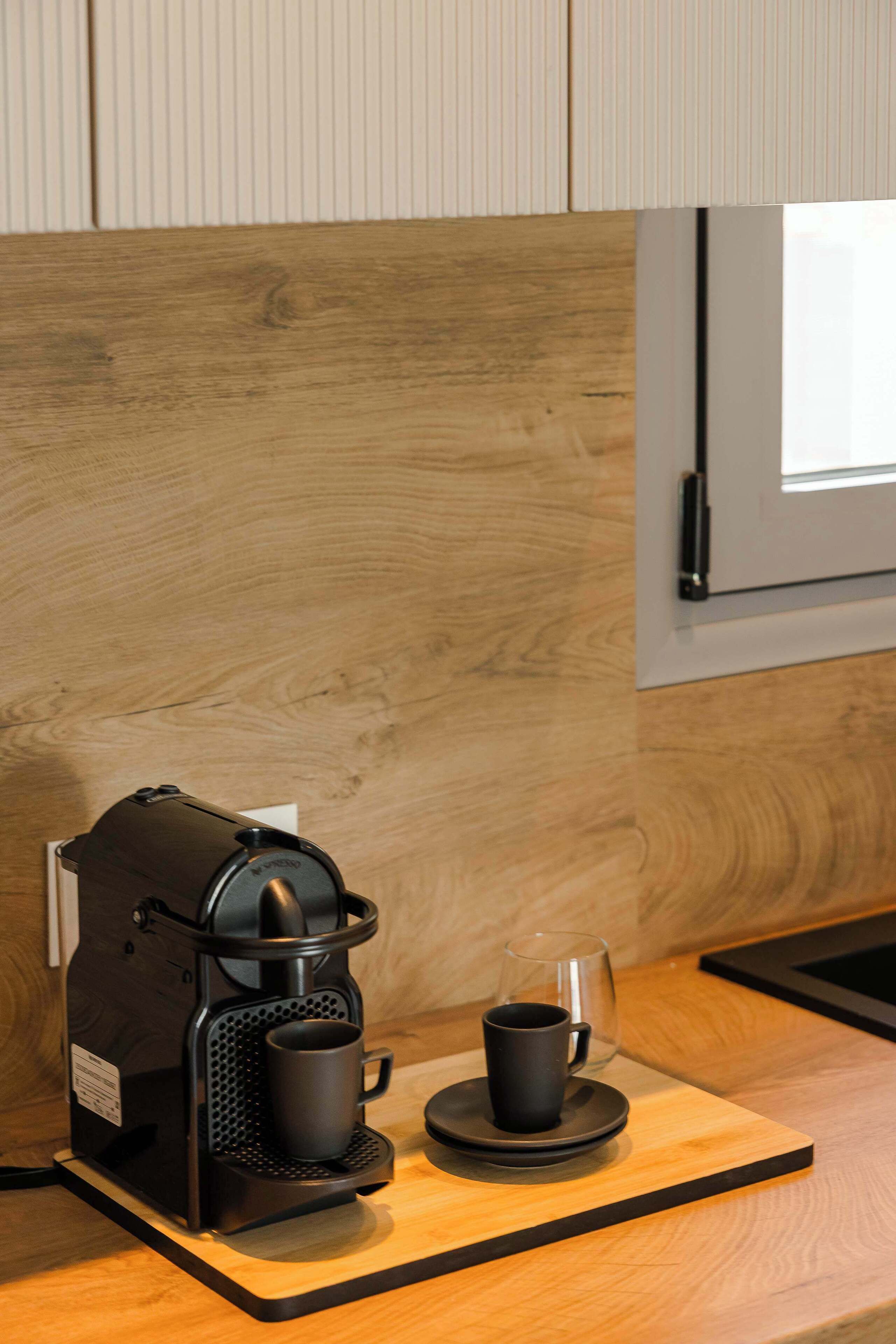 A compact coffee maker on a wooden countertop with a glass and a cup, against a backdrop of a wooden wall and a window.