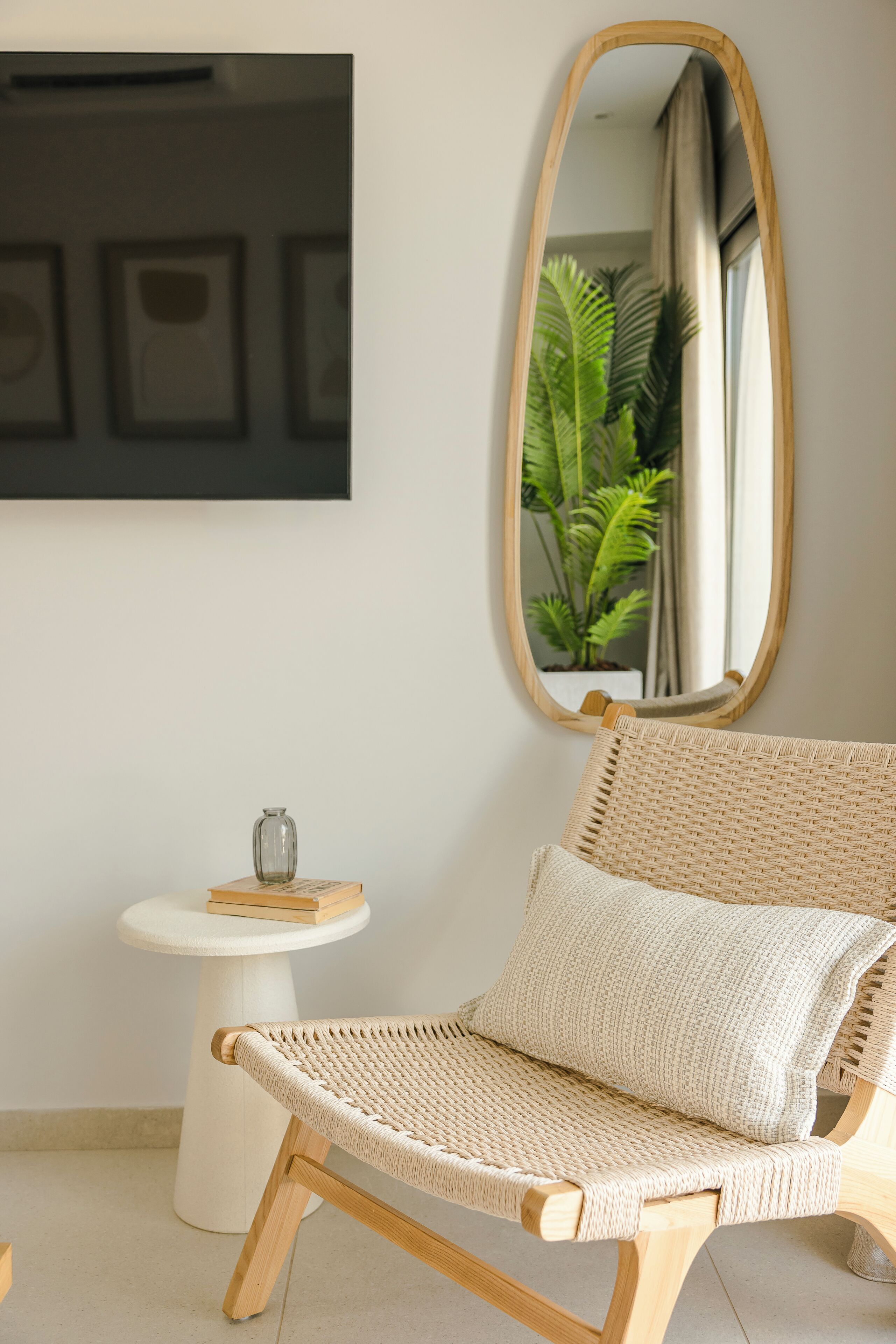 A wicker chair with a cushion, a small white table with a vase and a book, and a large mirror on the wall.