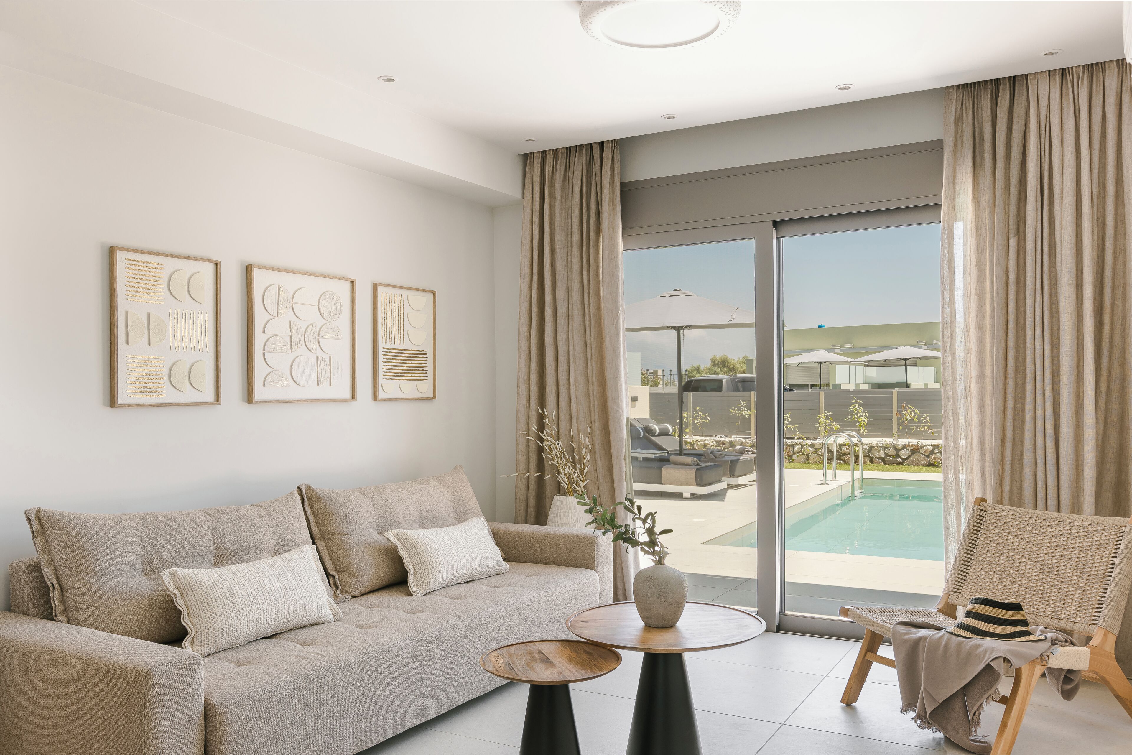 A modern living room with a sofa, a chair, a small table, and a large window with a view of a pool and outdoor furniture.