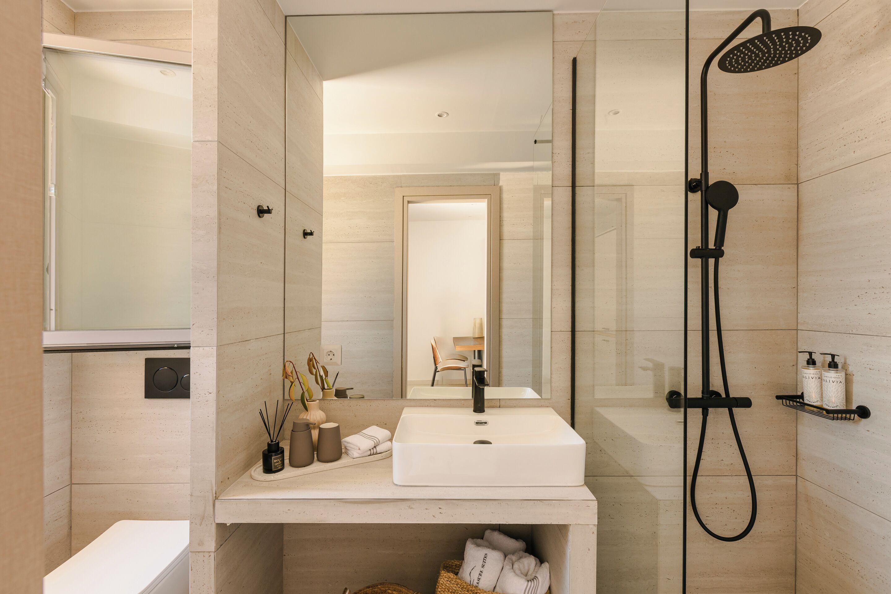 Un baño moderno con un espejo grande, un lavamanos blanco, un cabezal de ducha de mano y un perchero con artículos de aseo.