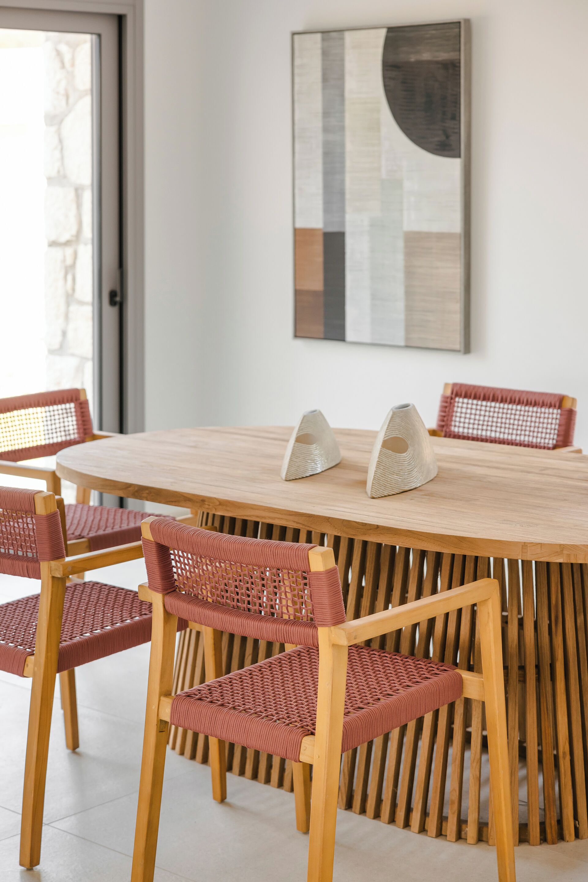 A round wooden table with six wicker chairs, a modern abstract painting on the wall, and a door with a stone wall visible through the glass.