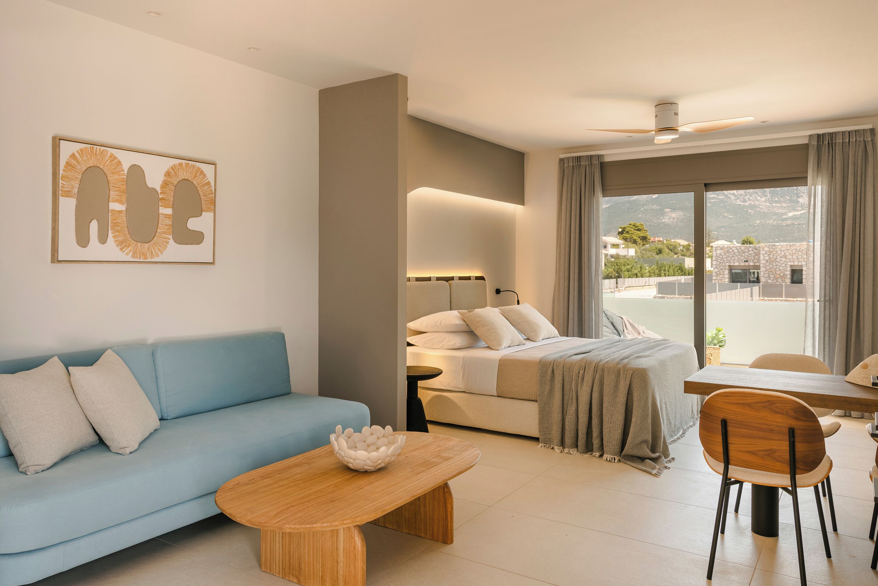 A modern hotel room with a sofa, a wooden coffee table, a bed with a nightstand, and a dining area with a table and chairs.