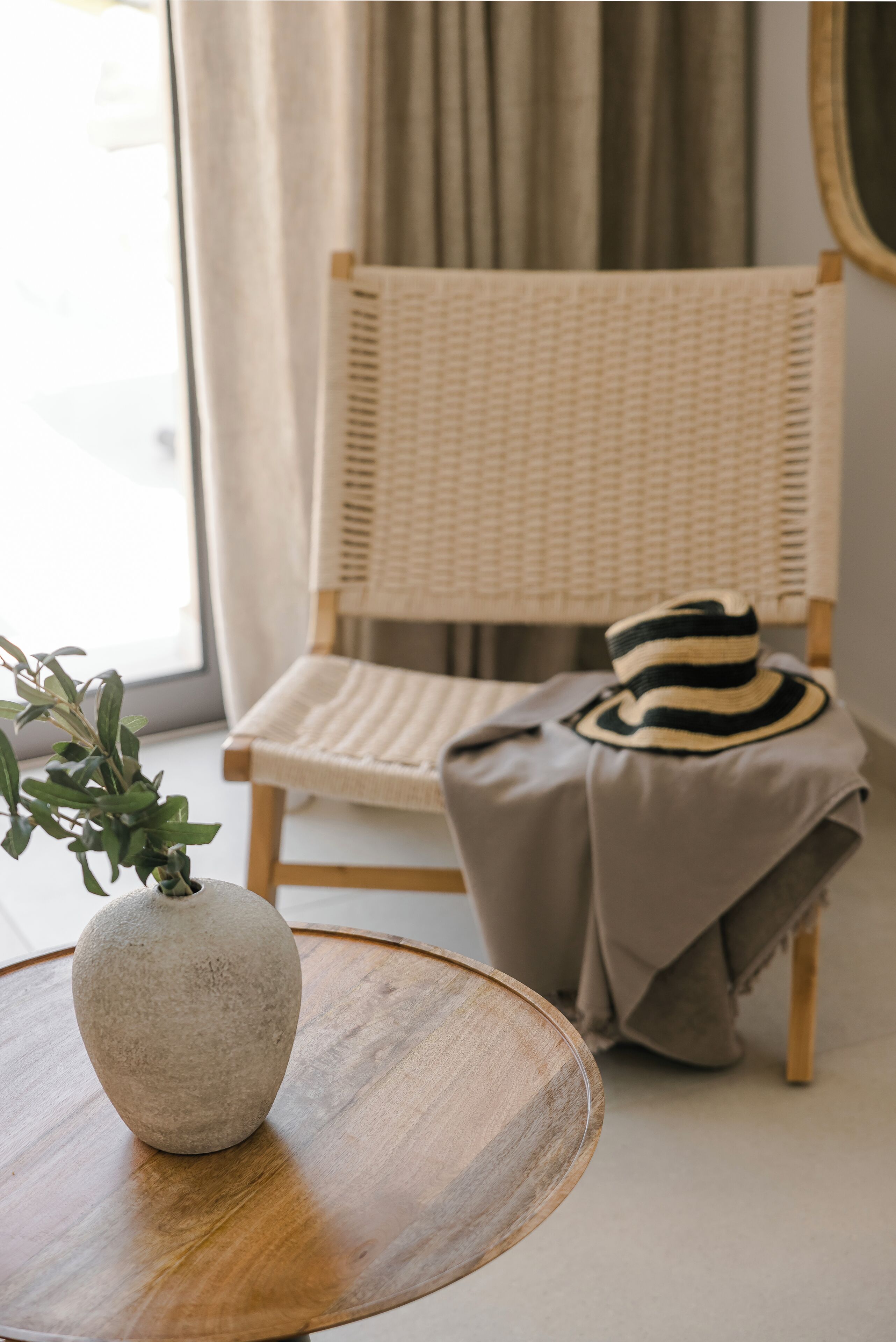 A wicker chair with a striped pillow, a round wooden table with a vase, and a plant.