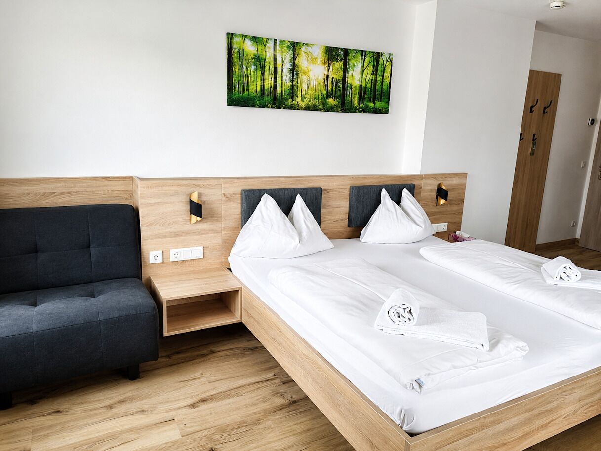 A modern hotel room with a wooden headboard, a grey sofa, a large bed with white linens, and a forest-themed wall art.