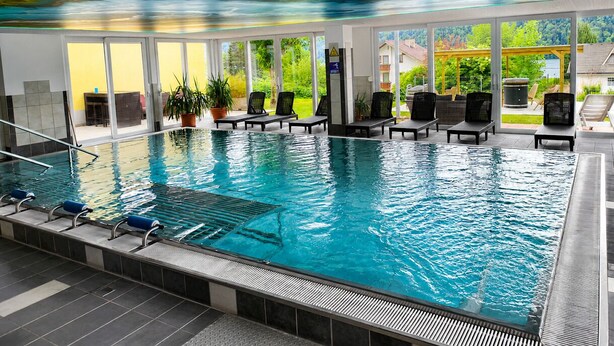 An indoor swimming pool with clear glass walls, lounge chairs, and a view of greenery outside. - Familienhotel Steindl (Millstatt)