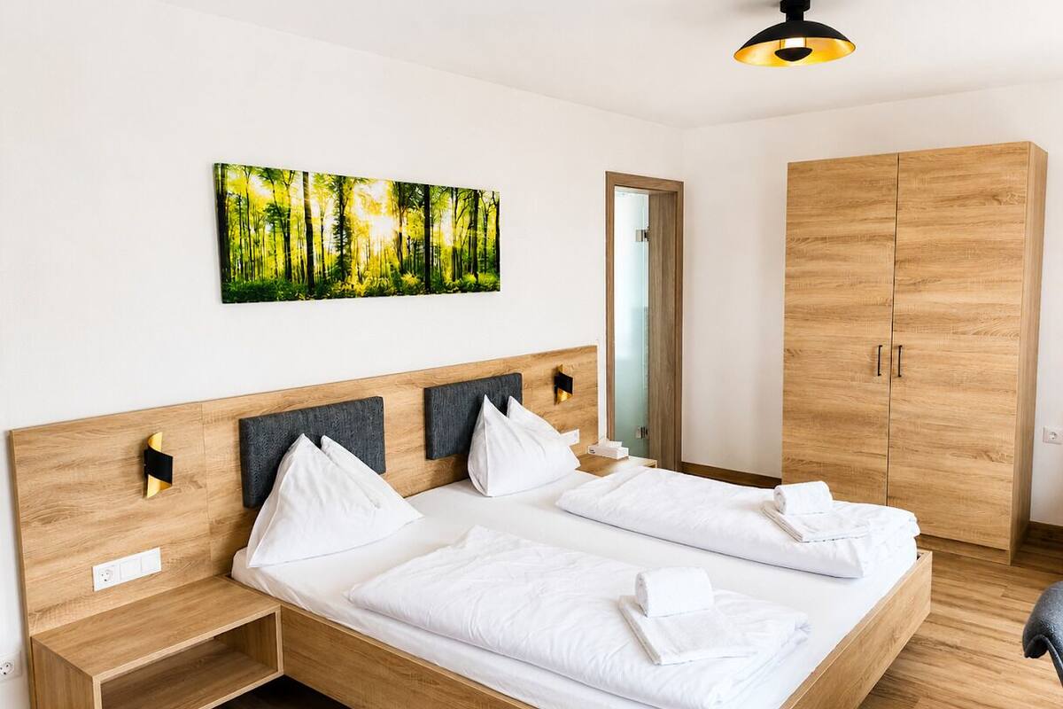 A double bed with white linens, a wooden headboard, a bedside table, a wardrobe, and a forest-themed wall art.