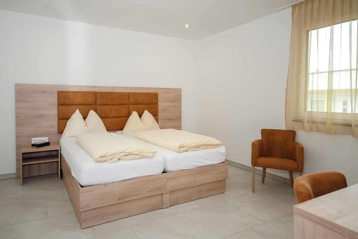 A modern bedroom with a wooden bed, a nightstand, a chair, and a window with sheer curtains.